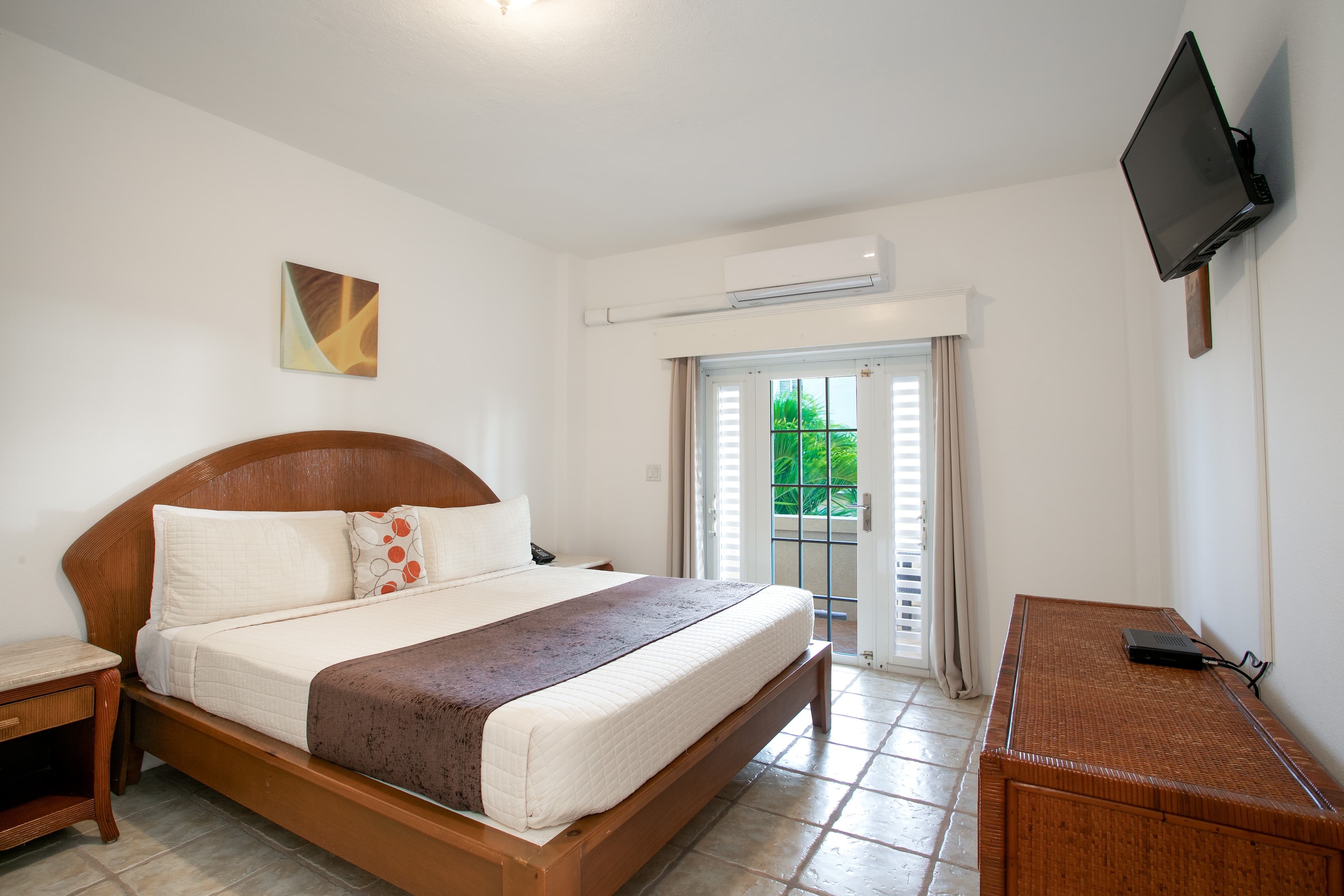 A neatly arranged hotel room featuring a large bed, a wooden dresser, and a TV, with natural light coming from a glass door.