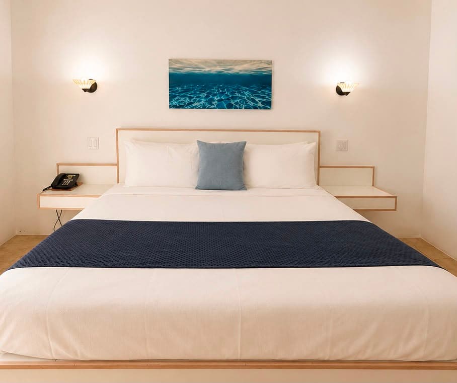 A minimalist bedroom featuring a large bed with white linens and a blue accent pillow, complemented by a water-themed artwork on the wall.