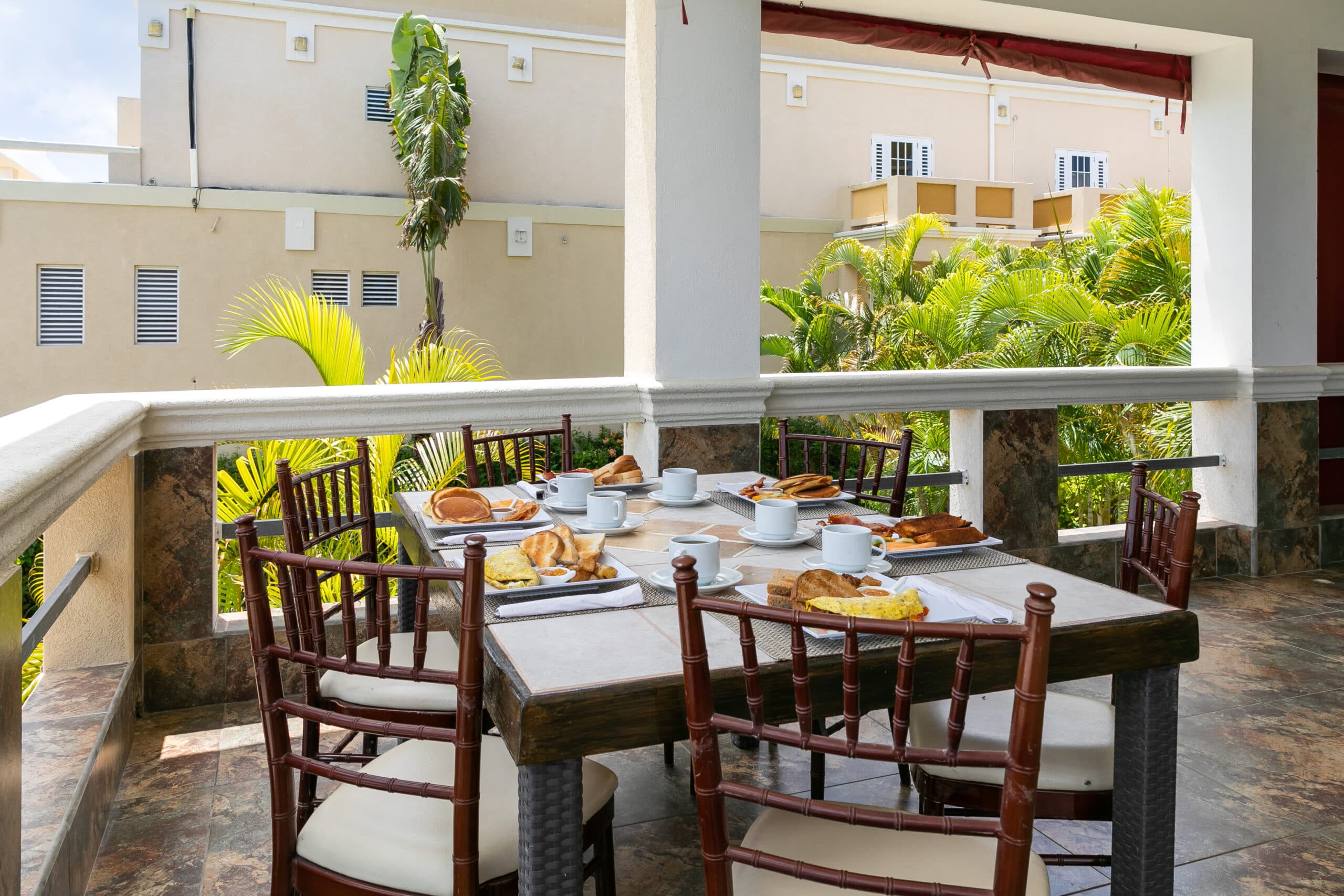 A breakfast table set with pastries and cups is surrounded by tropical greenery on a covered porch.