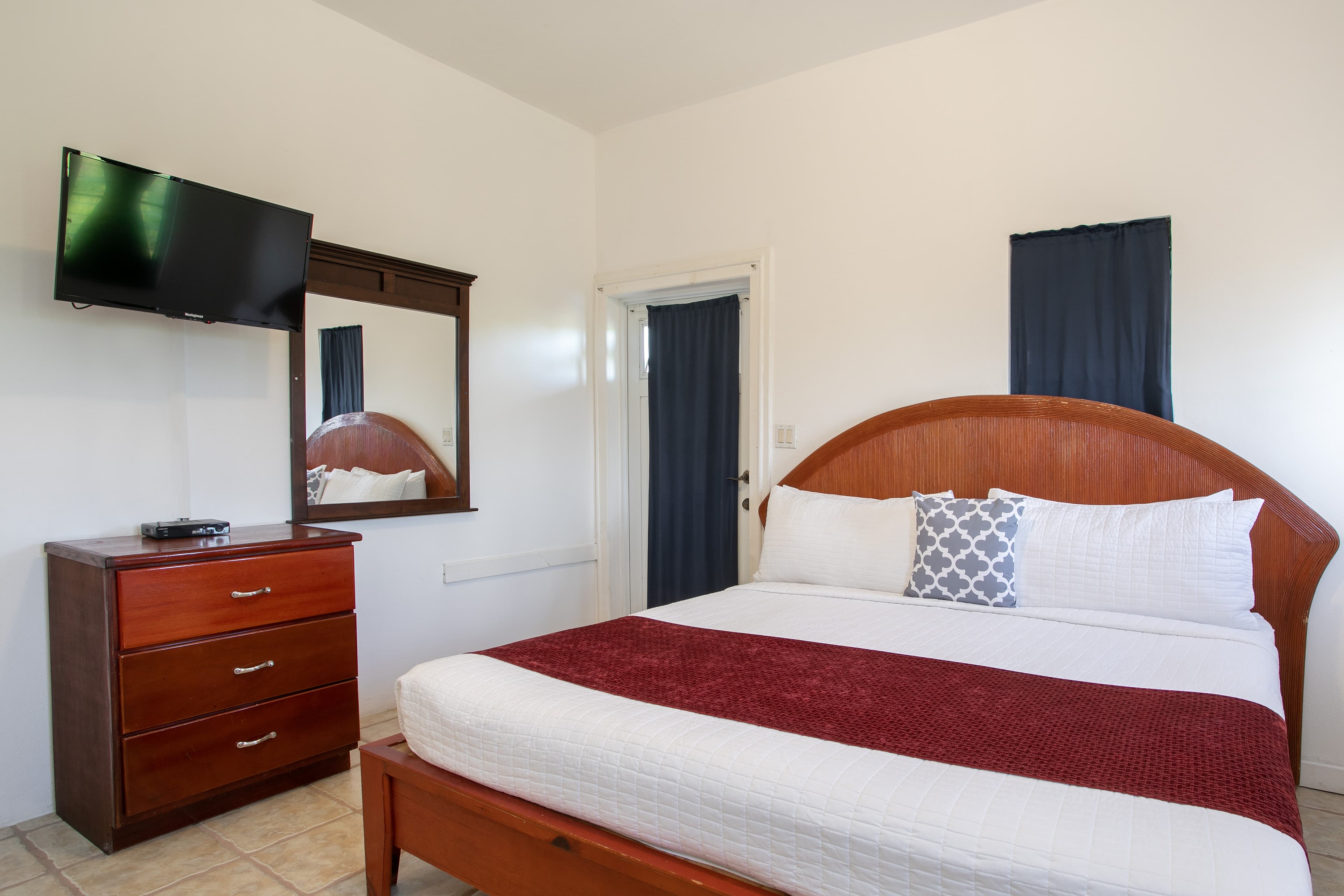 A cozy bedroom featuring a double bed with a red throw, a wooden dresser, a flat-screen TV, and a mirror.