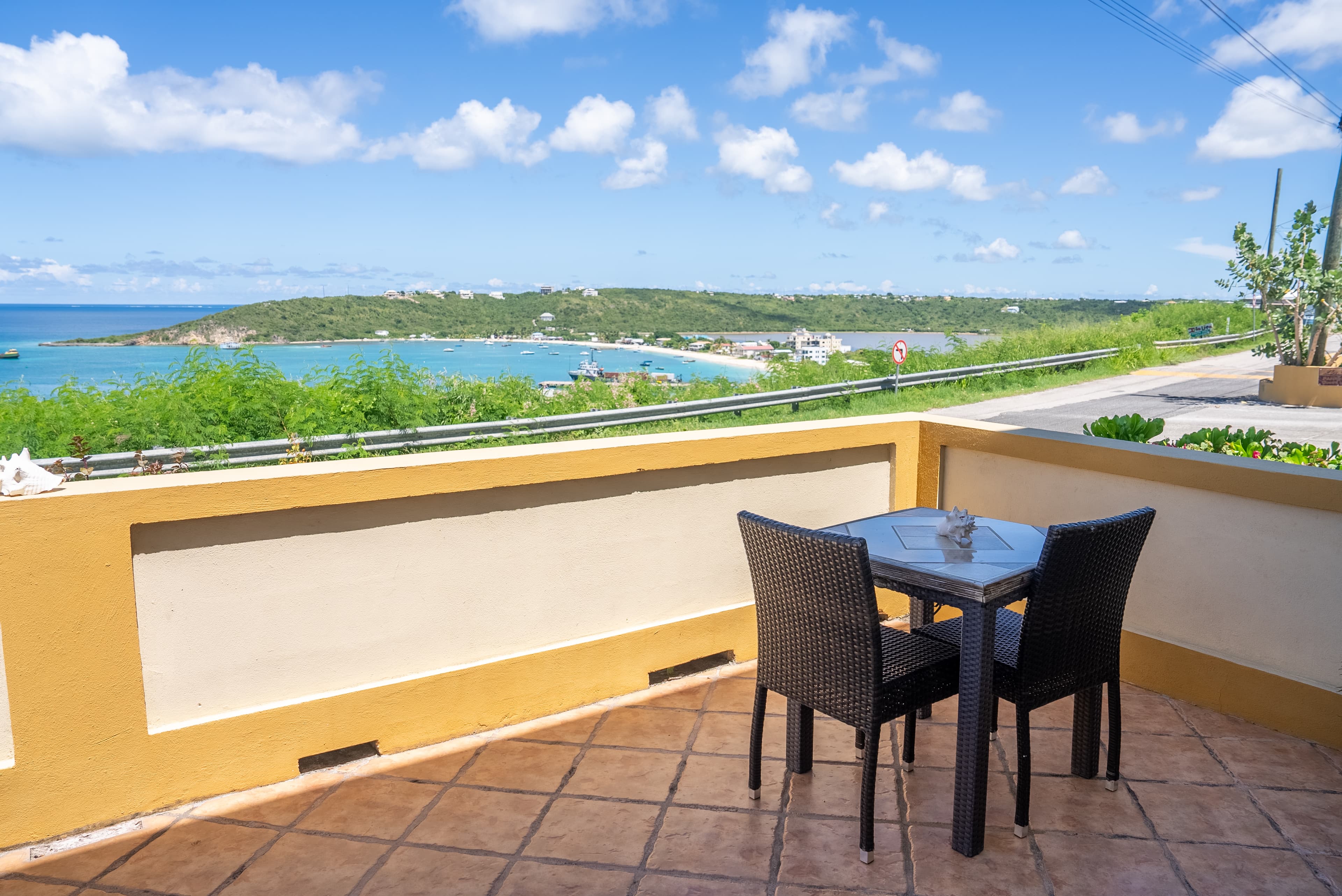 A small table and two chairs overlook a scenic coastal view under a blue sky with clouds.