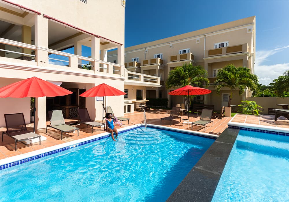 A sunny poolside scene featuring red umbrellas, lounge chairs, and a person relaxing by the water.
