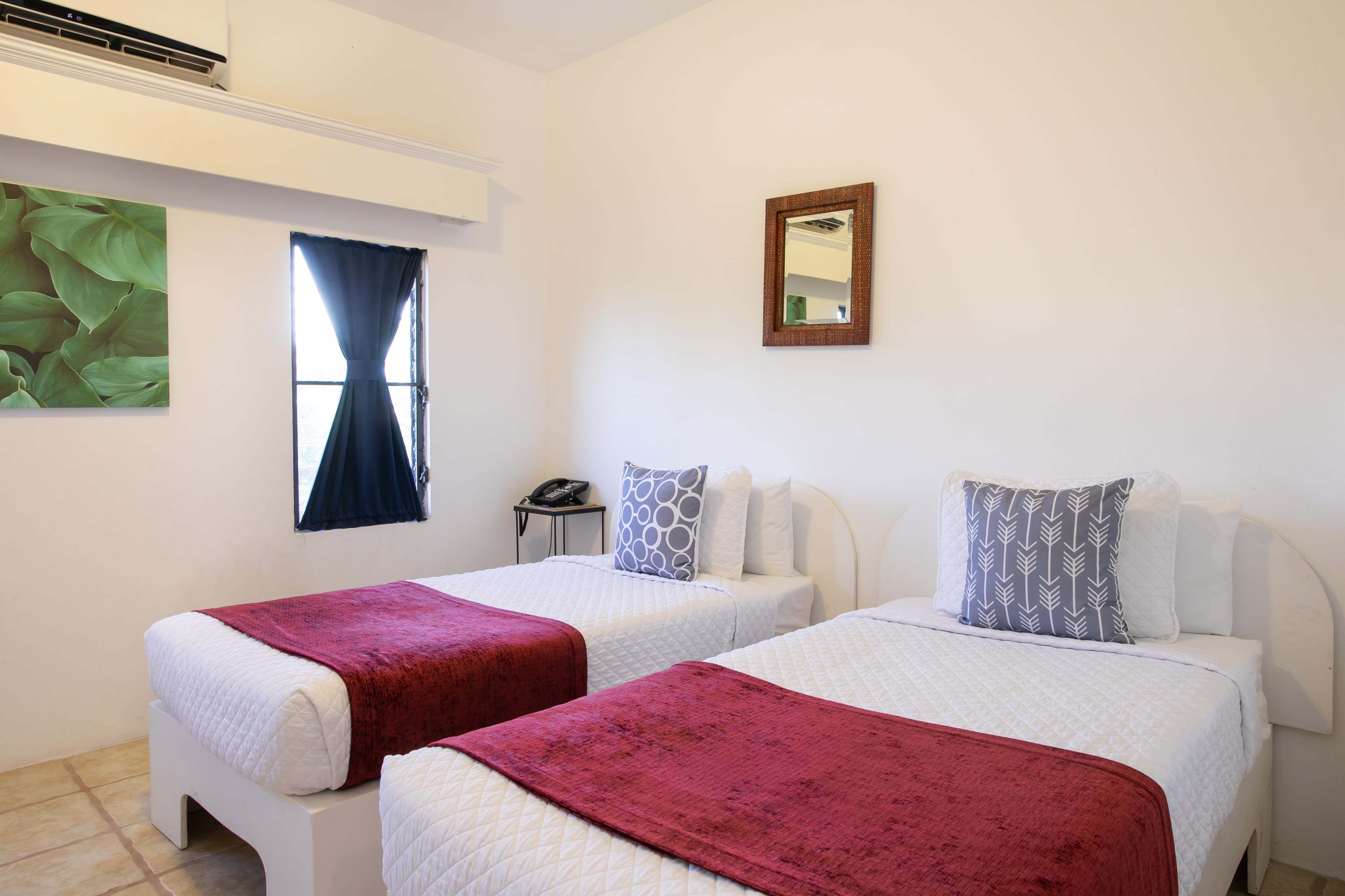 A well-lit hotel room featuring two twin beds with red throws, a mirror, and a window with dark curtains.