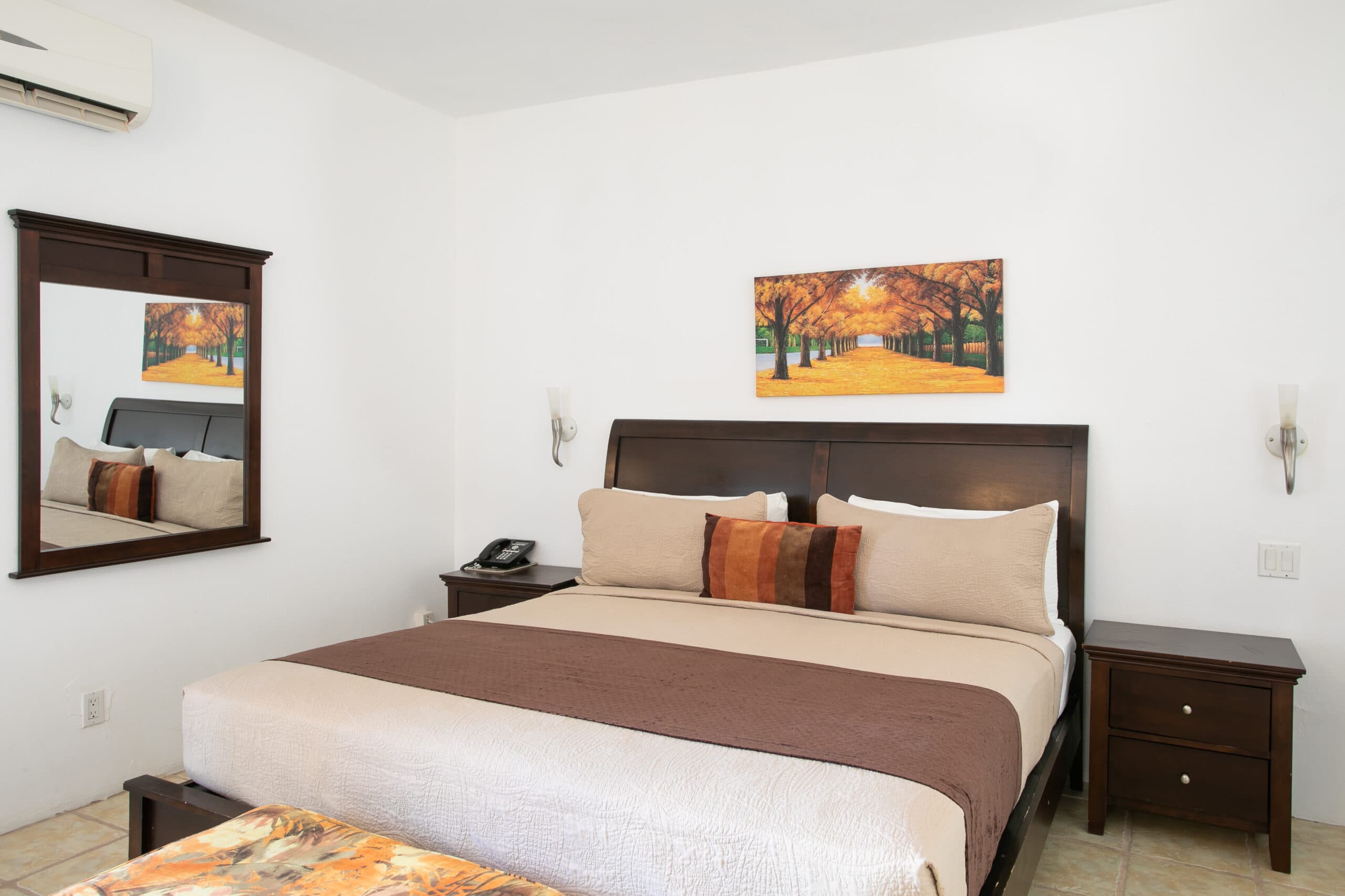 A neatly arranged bedroom featuring a large bed, a mirror, bedside tables, and an autumn-themed painting on the wall.