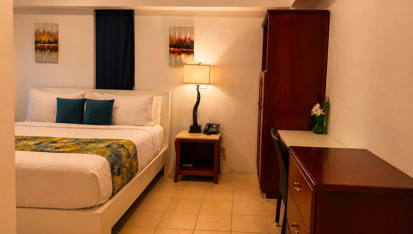 A cozy hotel room featuring a queen bed, desk, lamp, and wooden furniture.