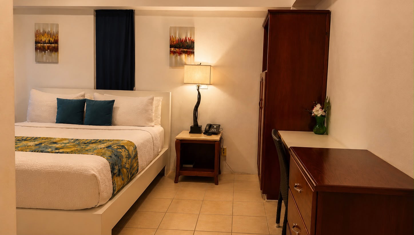 A cozy hotel room featuring a queen bed, desk, lamp, and wooden furniture.