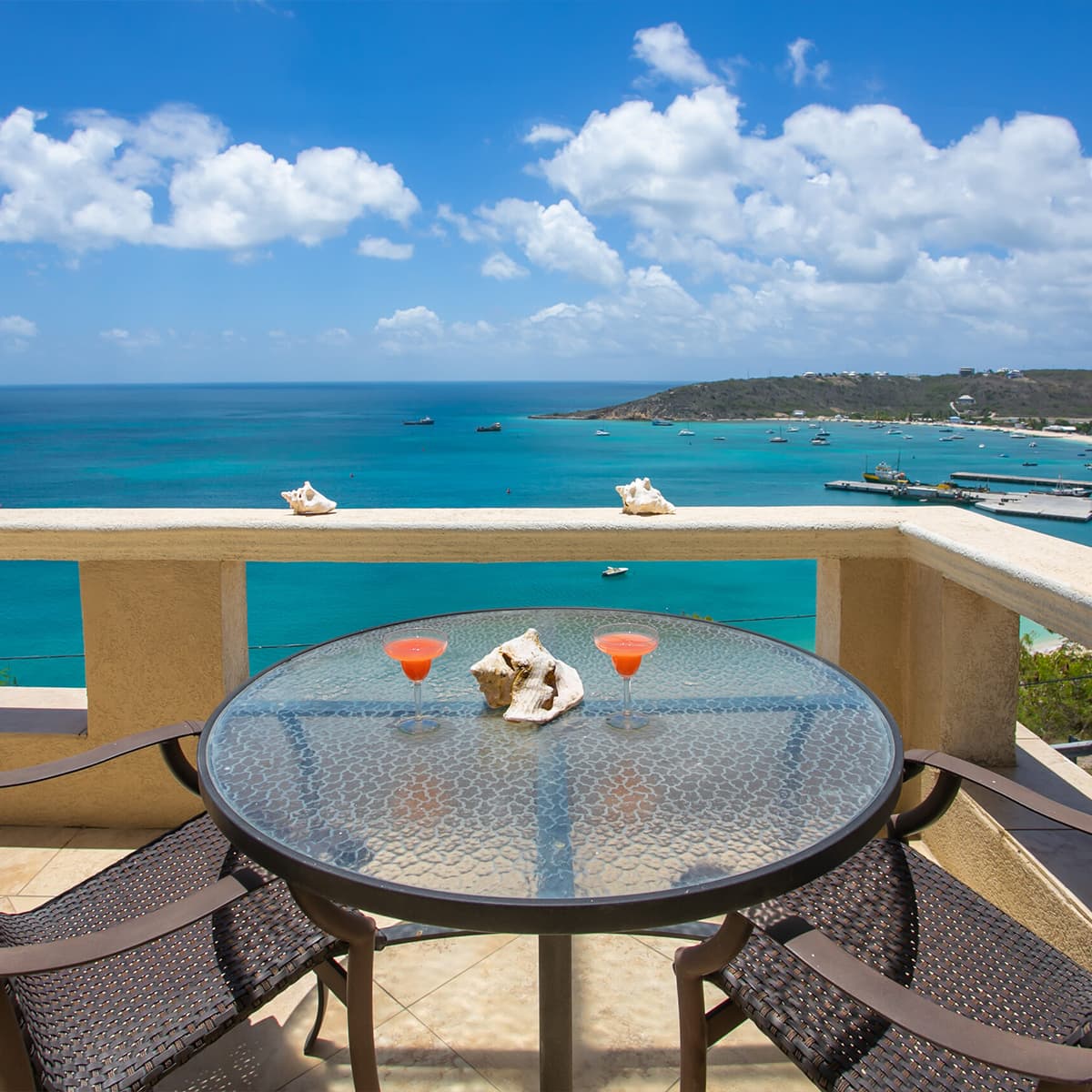 A terrace table set with two cocktails overlooks a scenic blue ocean and cloudy sky.