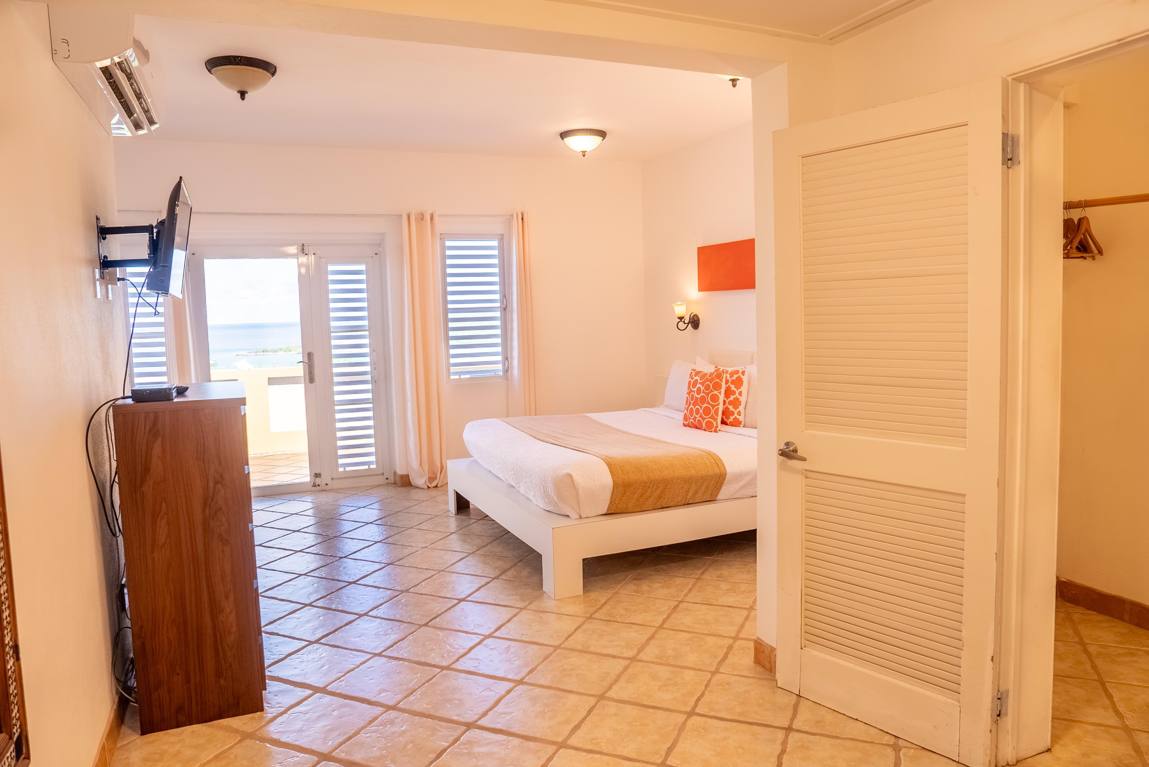 A bright hotel room featuring a bed with orange accent pillows, a TV, and access to a balcony through sliding glass doors.