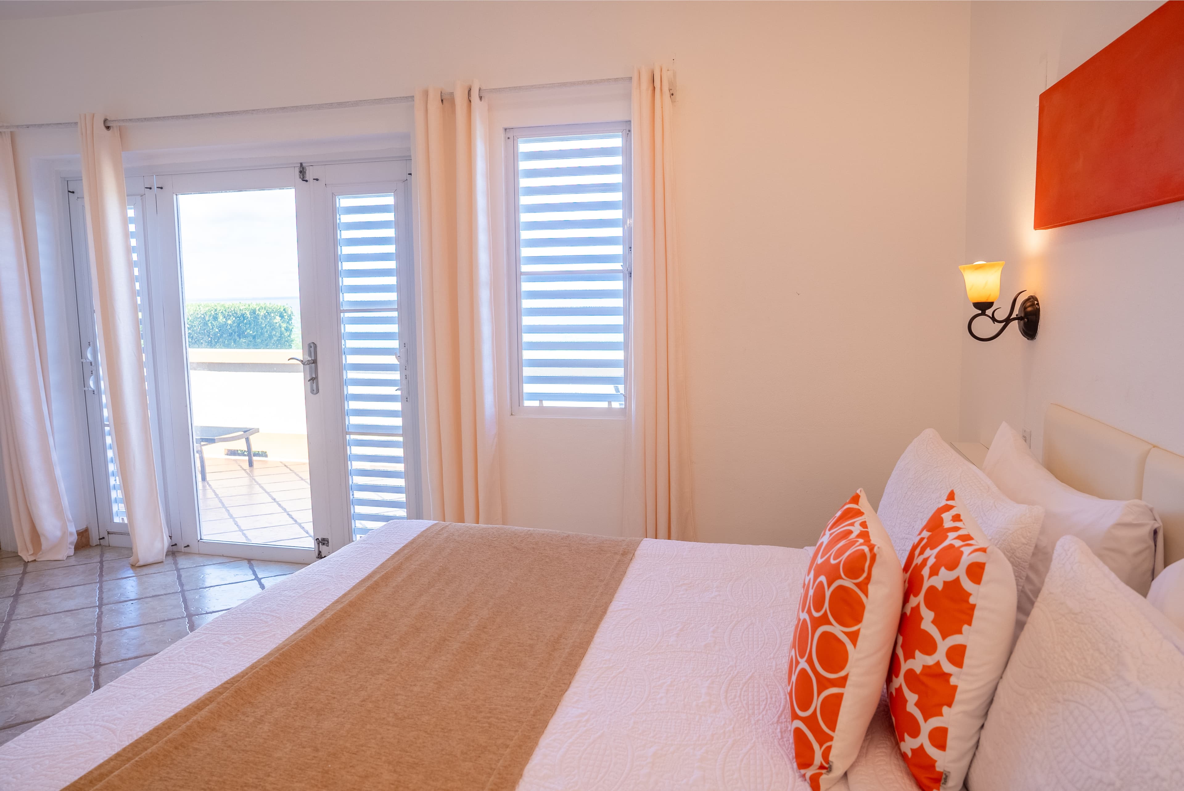 A cozy bedroom featuring a bed with orange patterned pillows, bright curtains, and a view of a balcony through sliding doors.