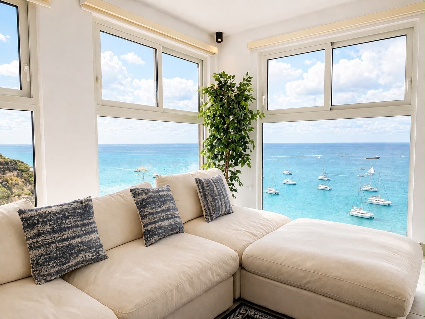 A bright living room features a cozy beige sofa with decorative pillows, framed by large windows overlooking a serene blue ocean and sailing boats.