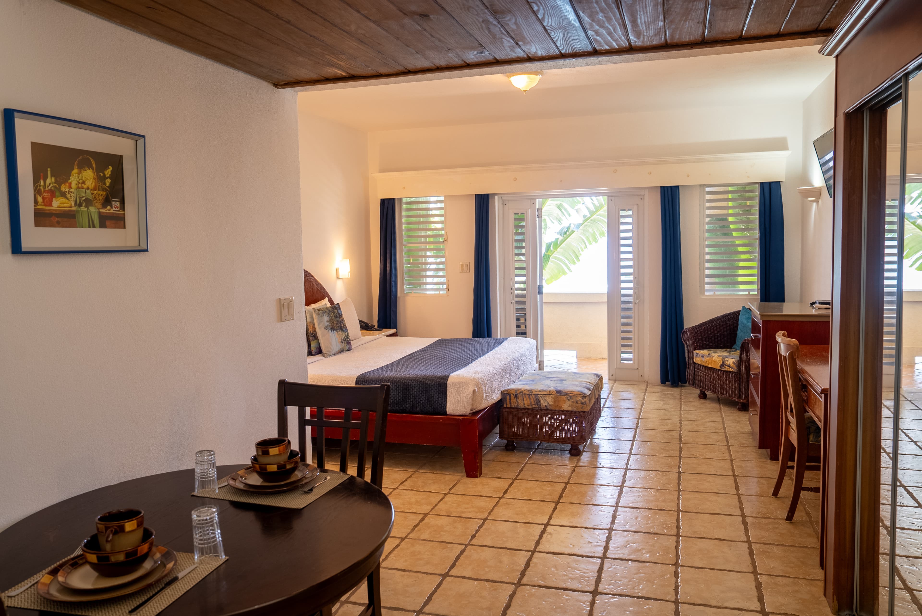 A bright, spacious hotel room featuring a bed, dining area, and large windows with blue curtains.