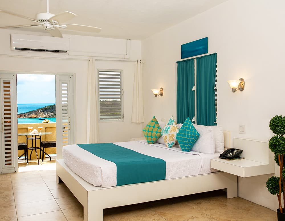 Bright hotel room with a large bed, turquoise accents, and a balcony overlooking the ocean.