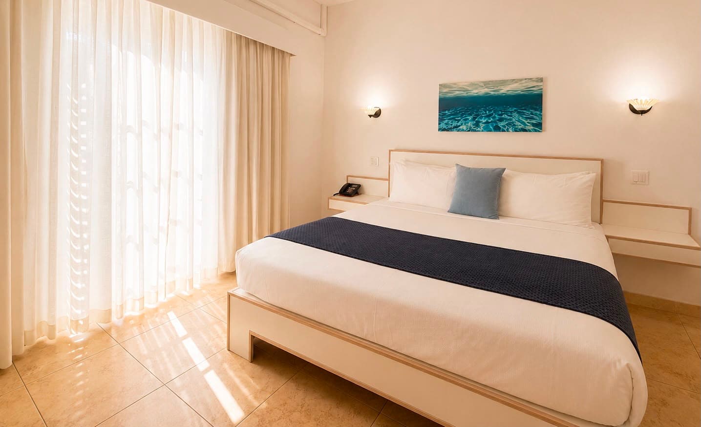 Bright and airy hotel room featuring a large bed, light-colored decor, and sheer curtains.