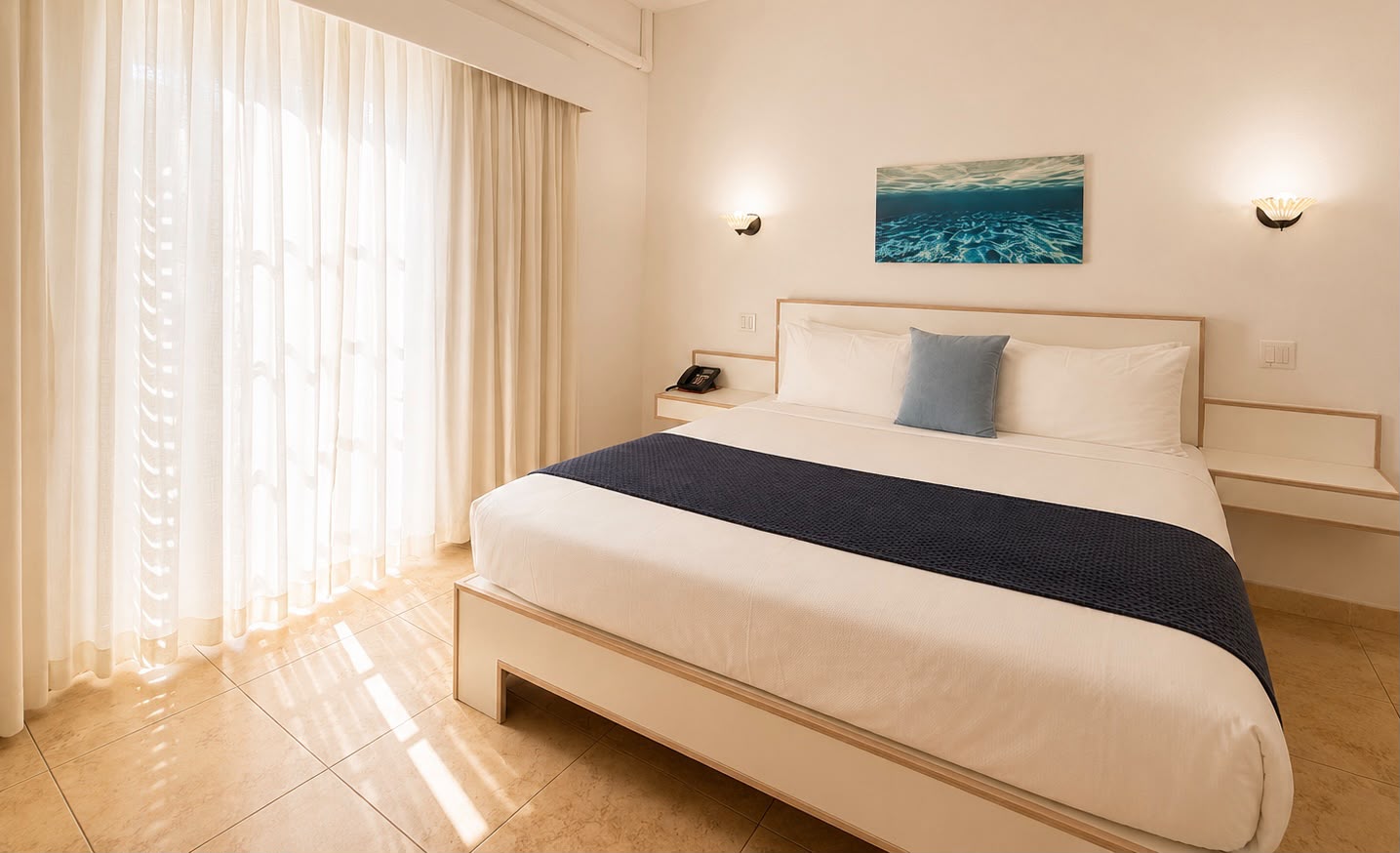 Bright and airy hotel room featuring a large bed, light-colored decor, and sheer curtains.