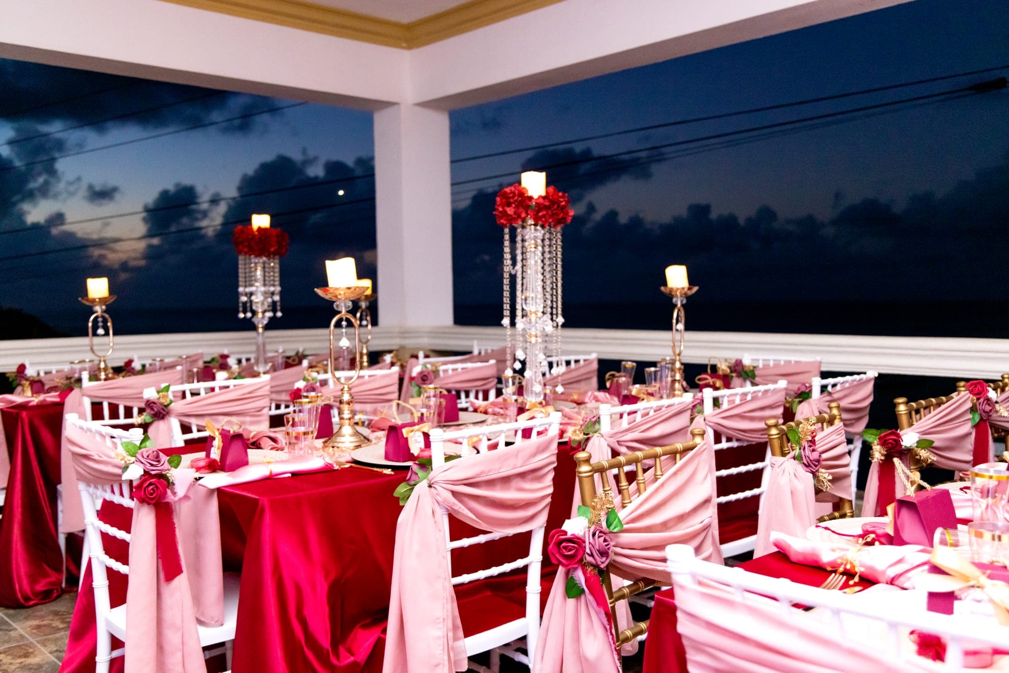 Elegantly decorated dining setup with pink and red accents, candles, and floral arrangements, overlooking a twilight sky.