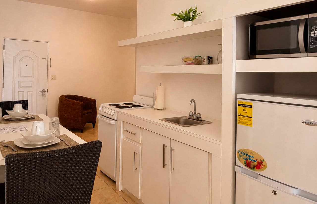 A modern kitchen featuring a stove, sink, refrigerator, and a small dining area with woven chairs.