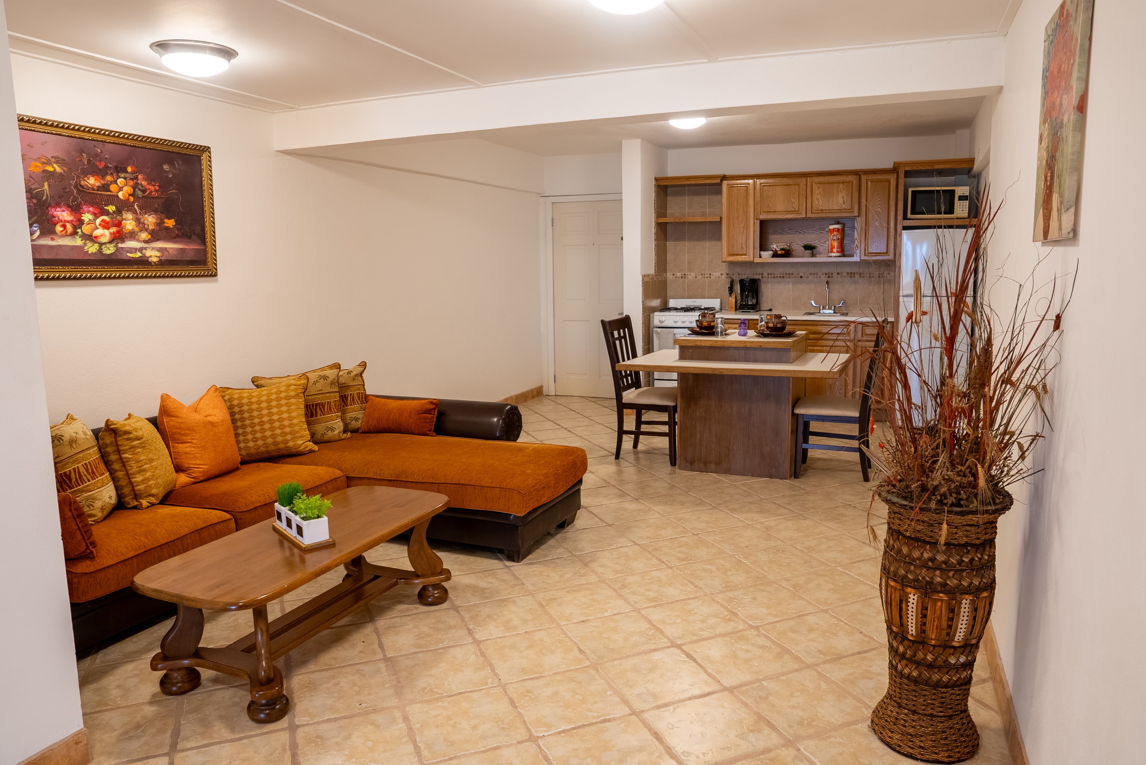 Cozy living area featuring an orange sectional sofa, a wooden coffee table, and a small dining space with a kitchen in the background.