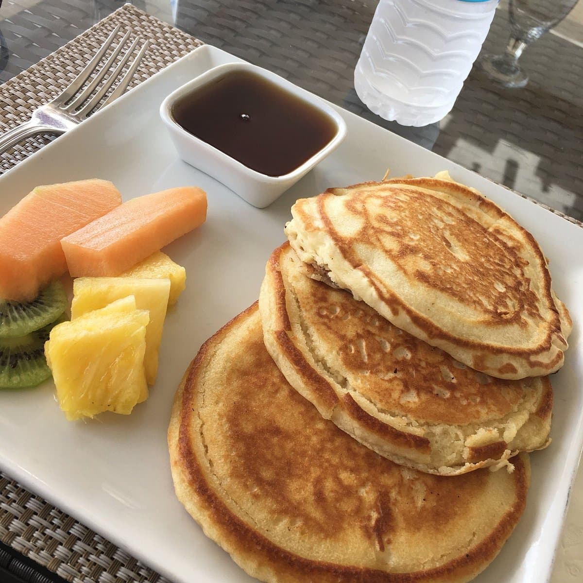 A plate of three golden pancakes with syrup and a side of fresh fruit including cantaloupe, pineapple, and kiwi.