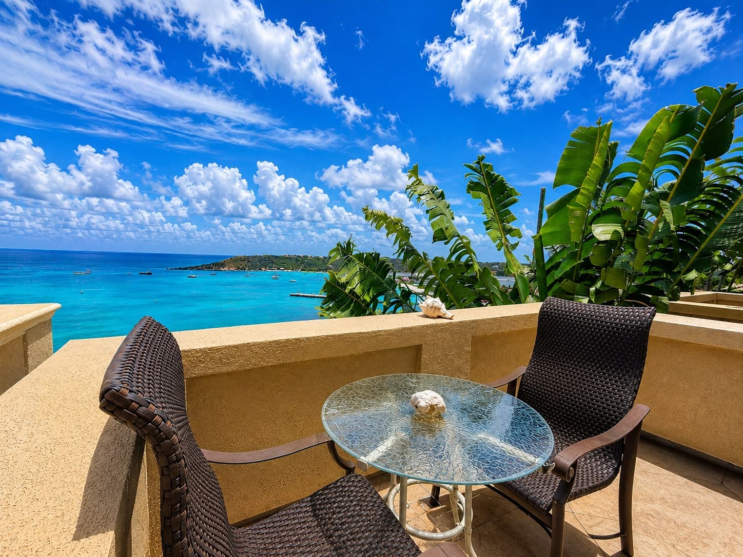 A coastal view of turquoise waters and lush greenery from a terrace with two chairs and a glass table.