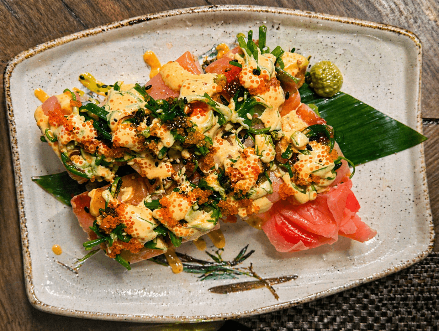 A decorative plate of sushi topped with green onions, orange roe, and drizzles of sauce, accompanied by wasabi and pickled ginger.