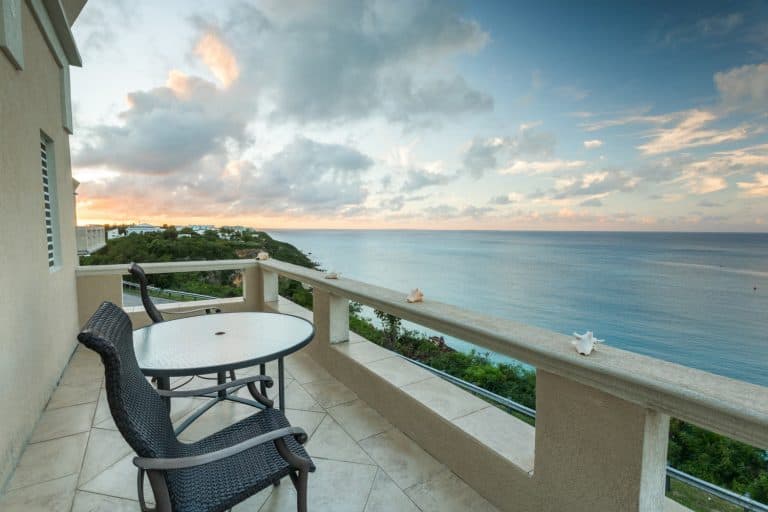 A balcony with a table and chairs overlooks a tranquil ocean at sunset.