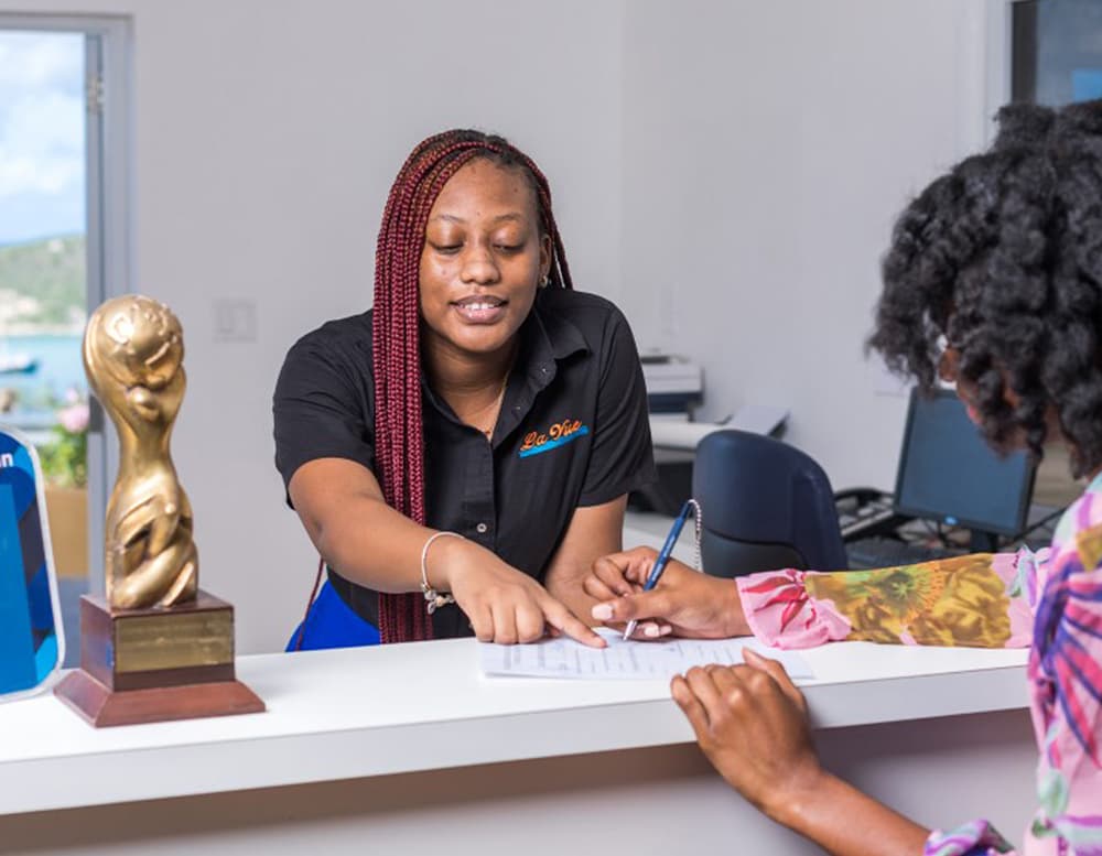 A receptionist assists a customer at a front desk, pointing at a document while an award statue is visible nearby.
