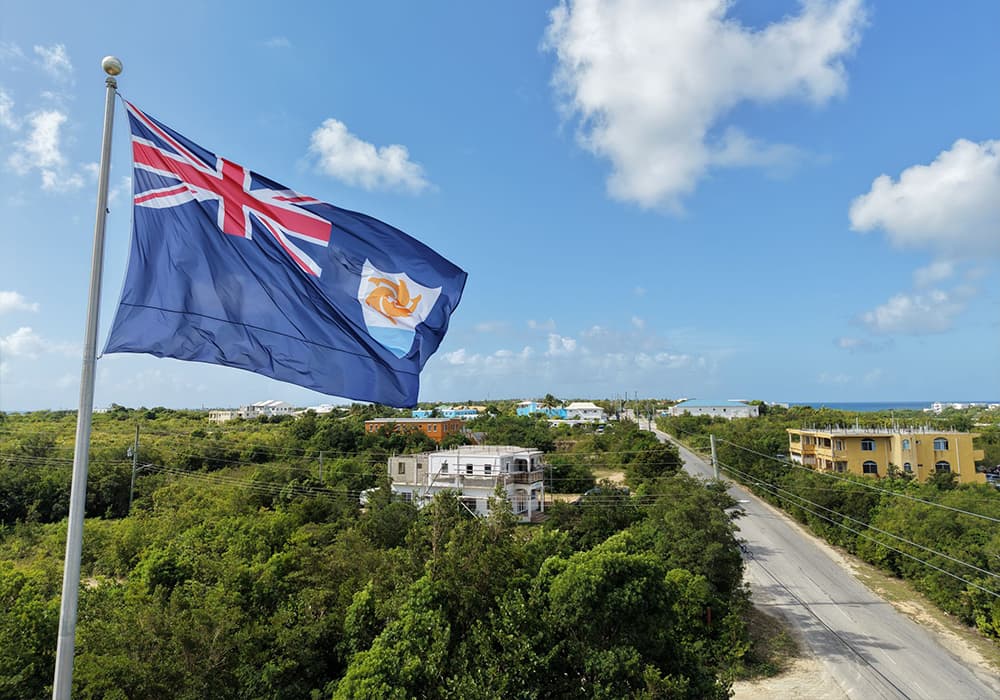 A flag with the Union Jack and a local emblem waves over a lush, green landscape and a winding road in Anguilla.