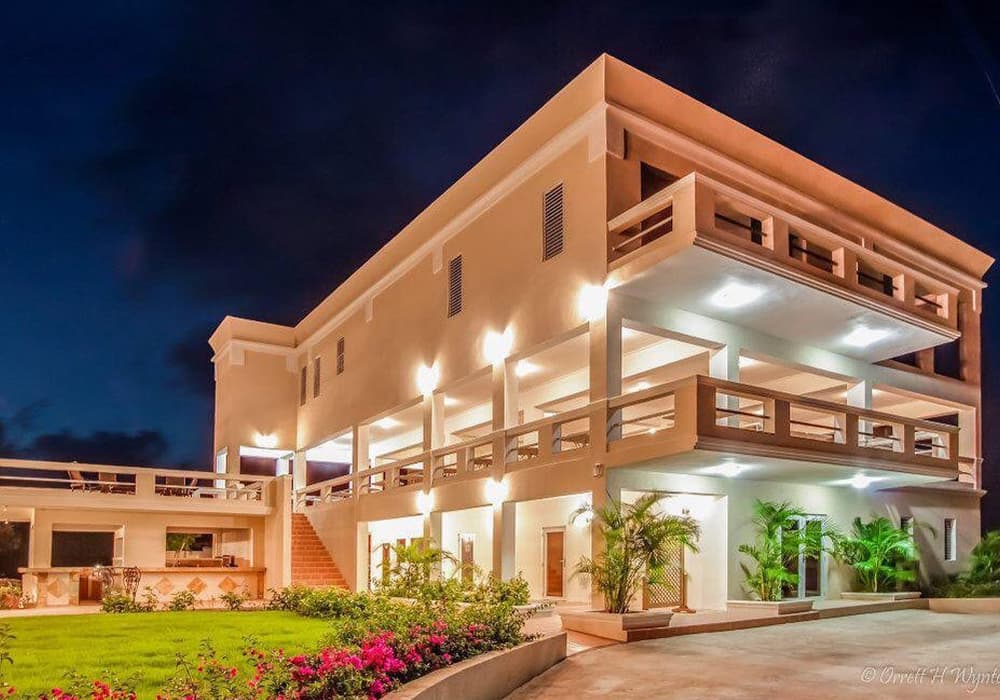A modern, multi-level house illuminated at night, surrounded by lush greenery and flowers.