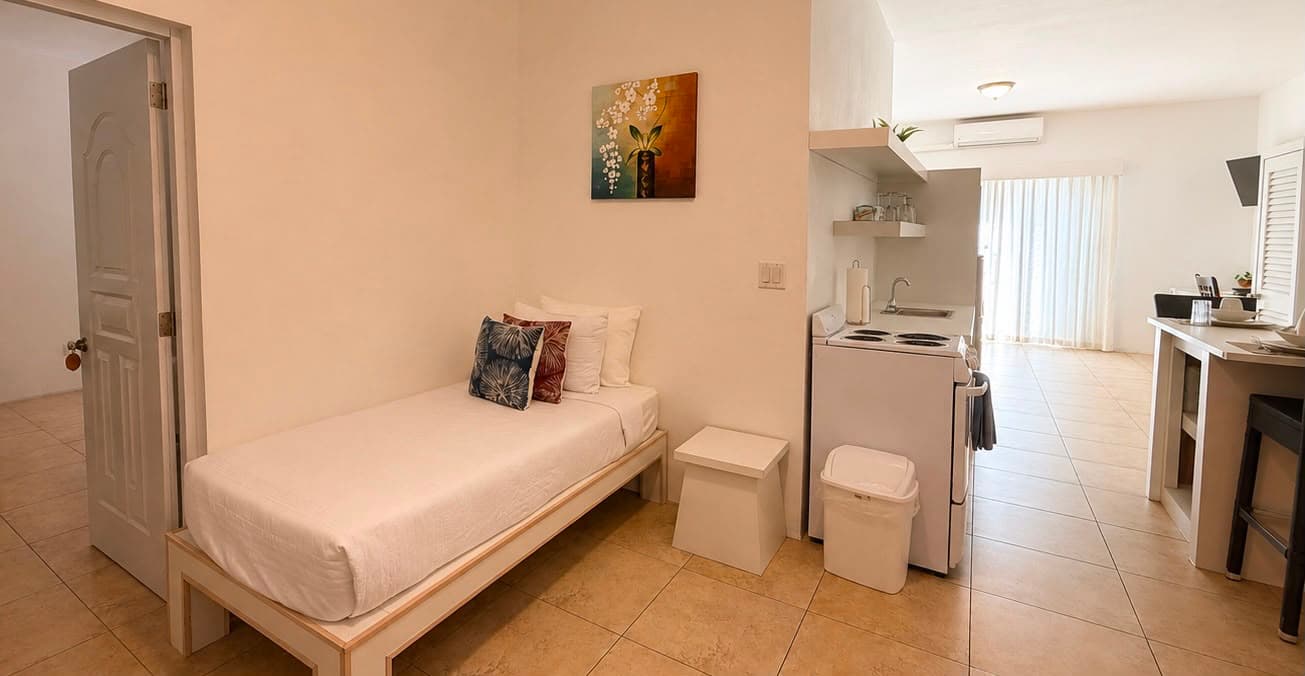 A cozy, minimalist bedroom with a single bed, a small kitchenette, and light-colored tile flooring.