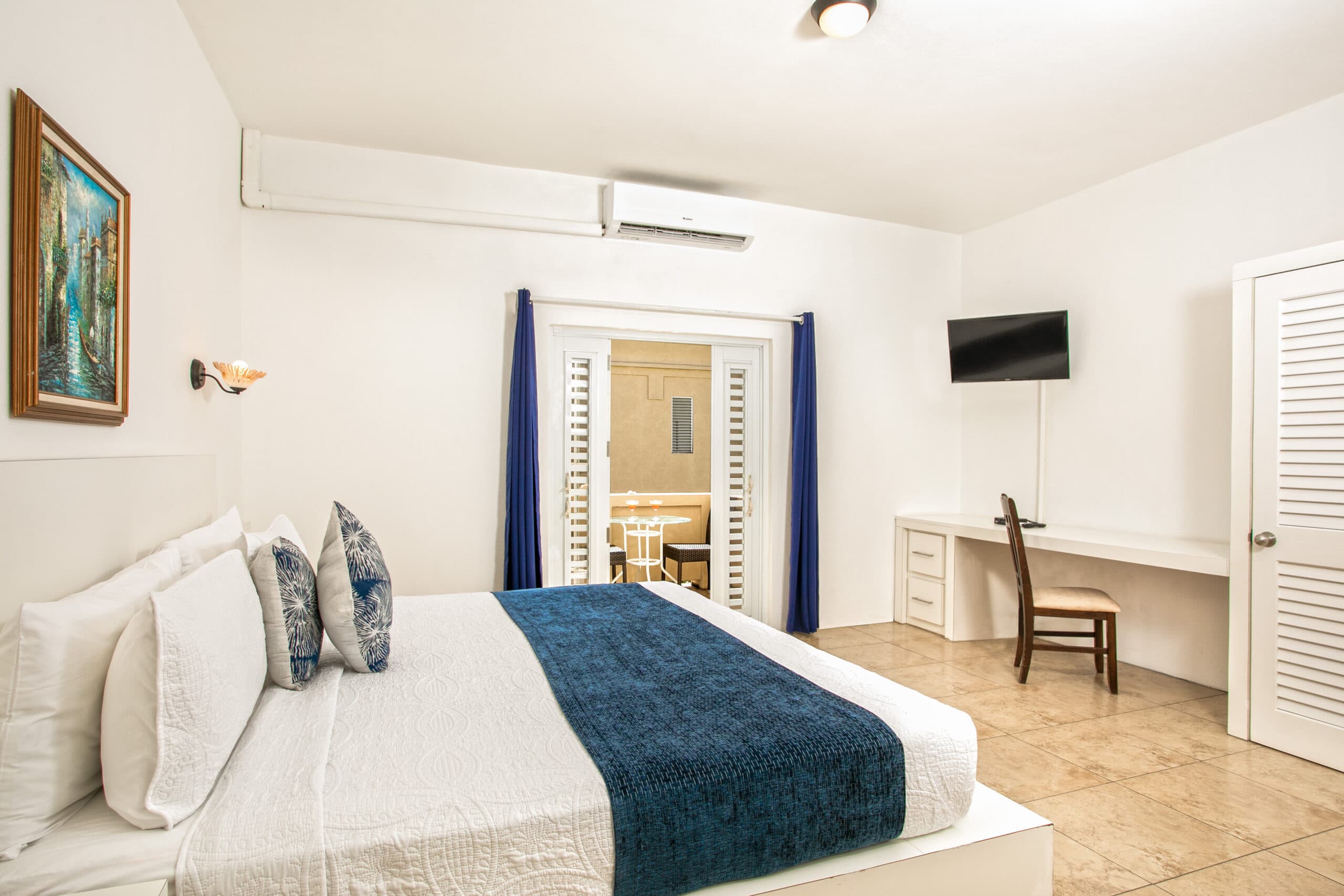 A bright, modern bedroom featuring a large bed with blue and white accents, a desk, chair, and a wall-mounted TV.