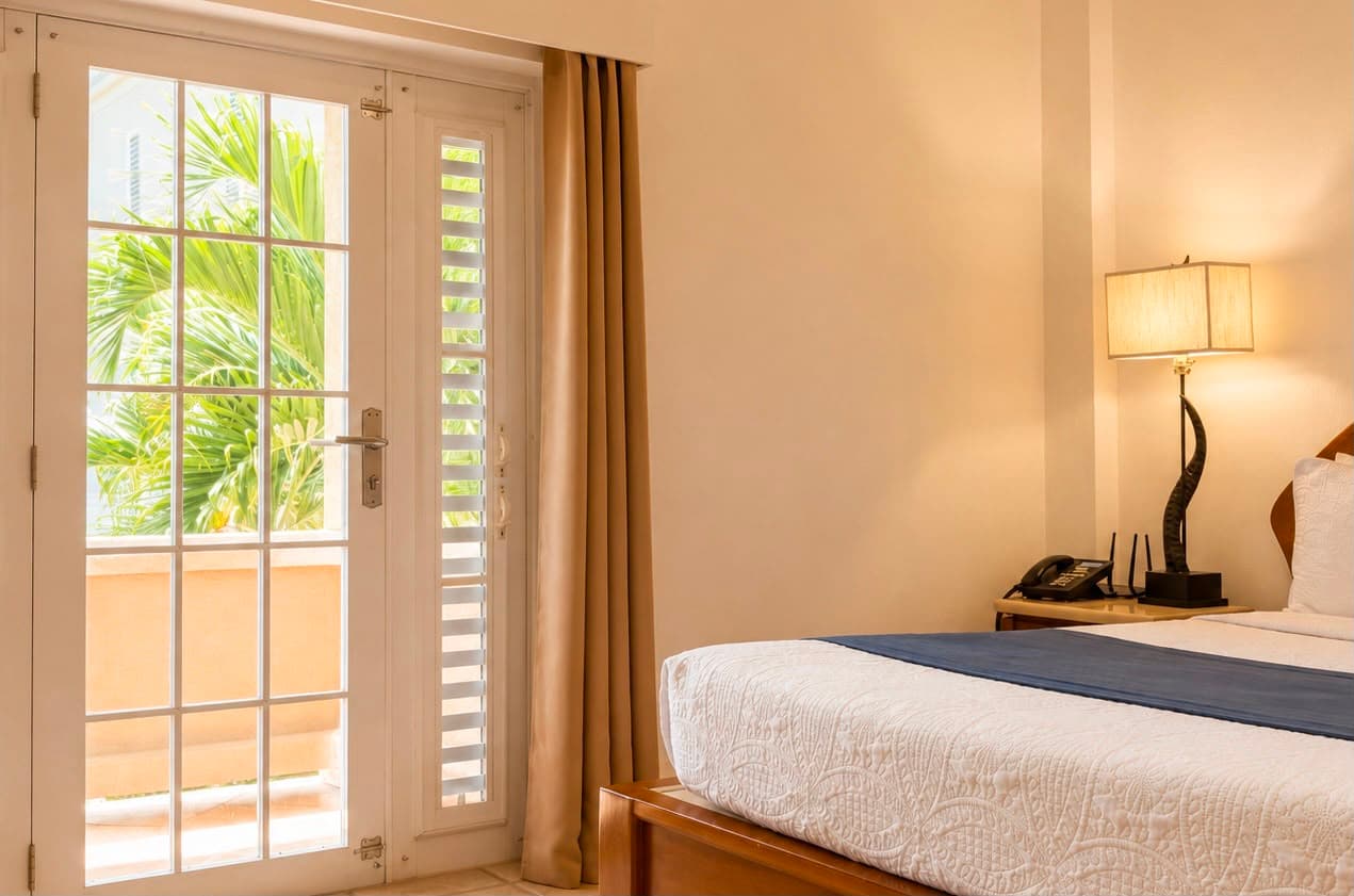A bright hotel room featuring a bed, lamp, and French doors leading to a balcony with greenery visible outside.